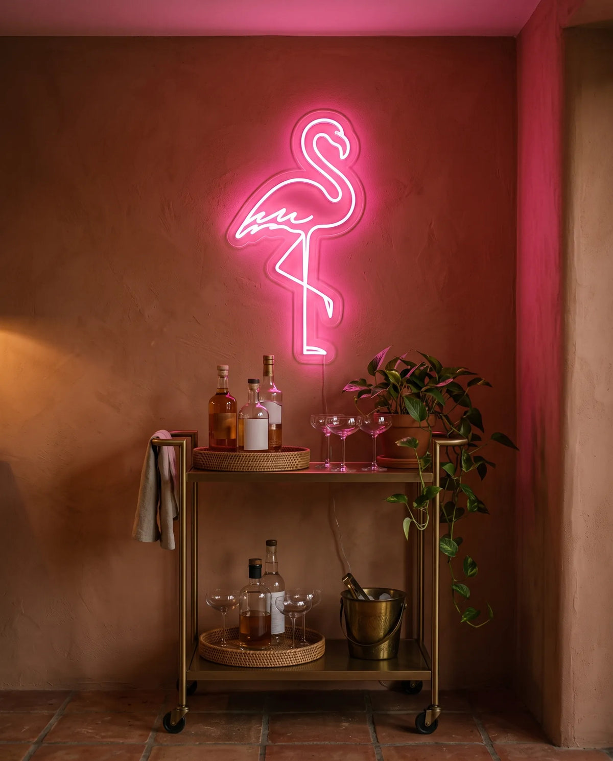 Néon LED flamant rose allumé au-dessus d’une desserte de bar en laiton, coupes et bouteilles sur mur terracotta