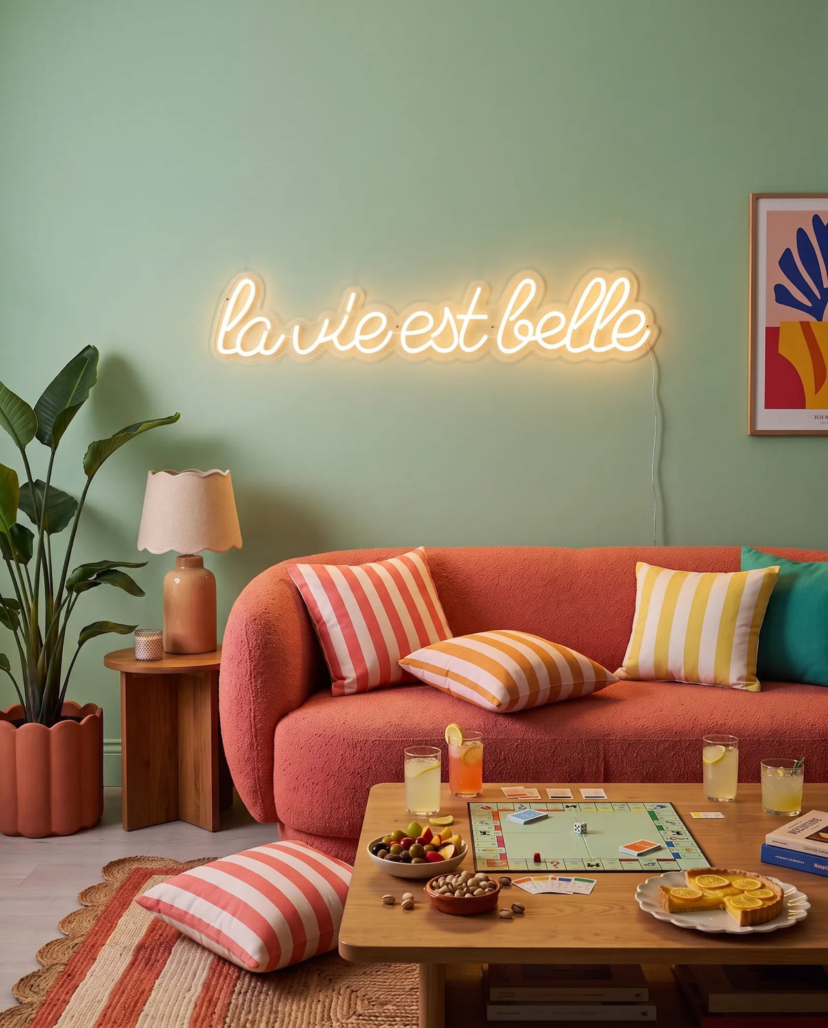Néon LED citation la vie est belle sur un mur vert d’eau, canapé corail, coussins rayés et limonades
