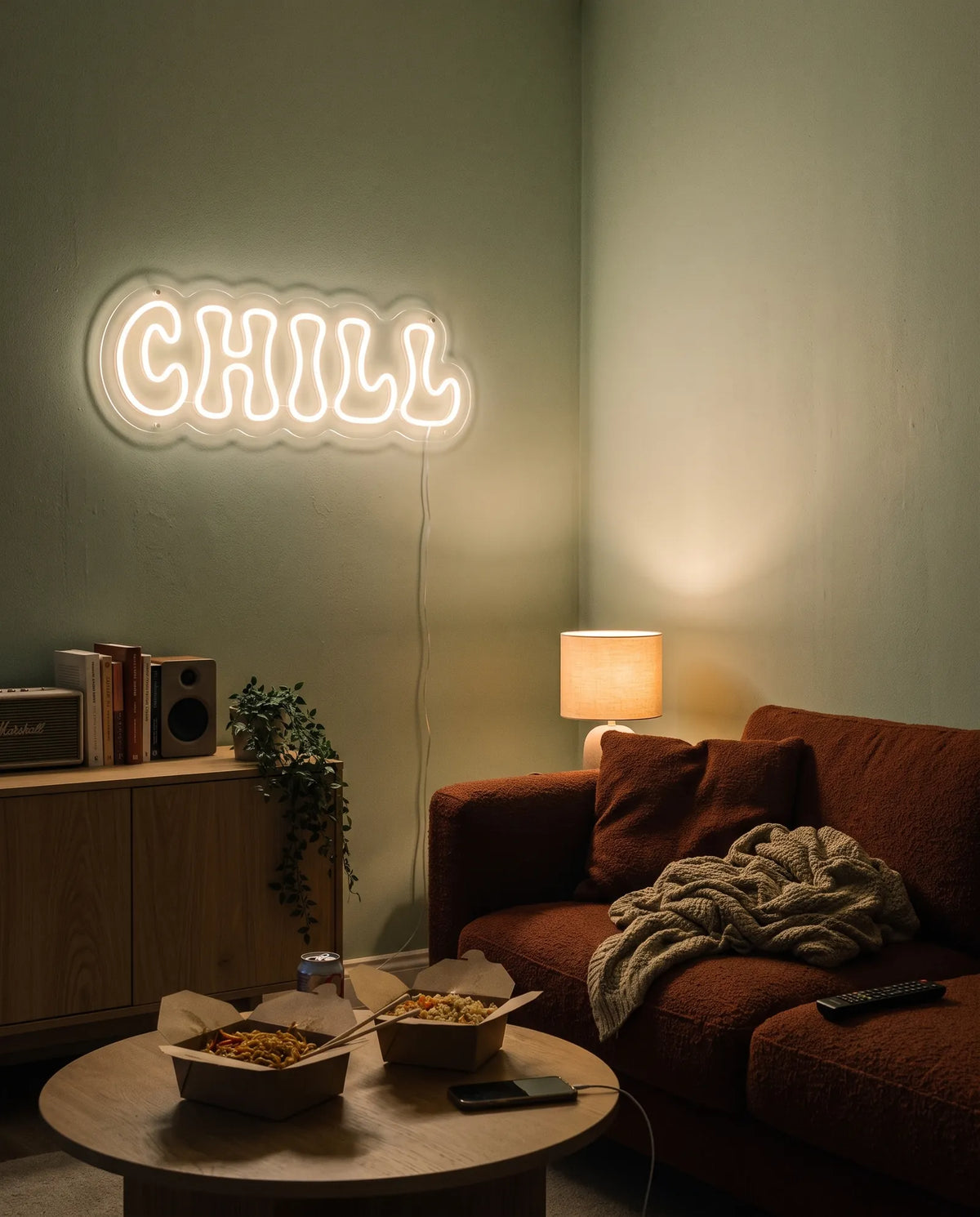 Néon chill blanc chaud allumé dans un salon, canapé rouille, plaid et plats à emporter sur la table basse