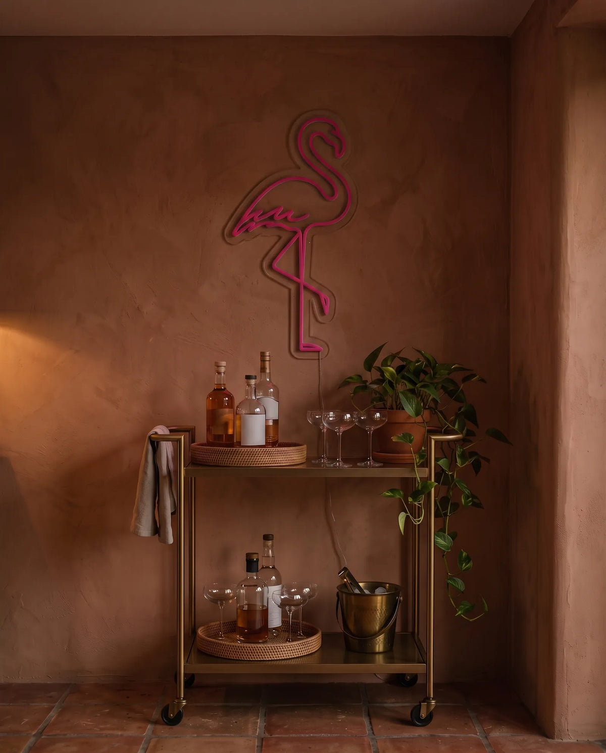 Néon flamant rose éteint au-dessus d’une desserte de bar dorée, mur terracotta en fin de journée