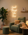 Néon Good Things Take Time blanc chaud allumé au-dessus d’un fauteuil vert, pouf, plaid et livre ouvert