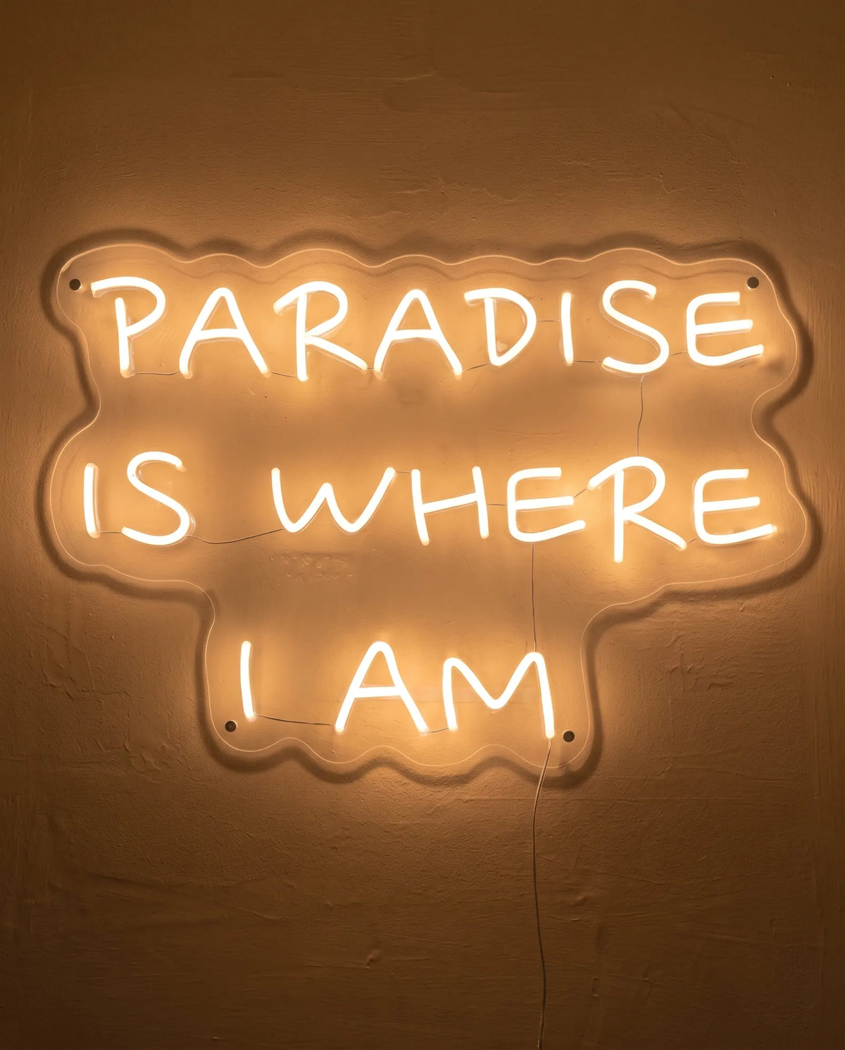 Néon Paradise Is Where I Am de face sur mur peint, capitales manuscrites sur trois lignes