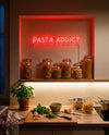 Néon Pasta Addict rouge allumé au-dessus de bocaux de pâtes, basilic, parmesan et planche à découper
