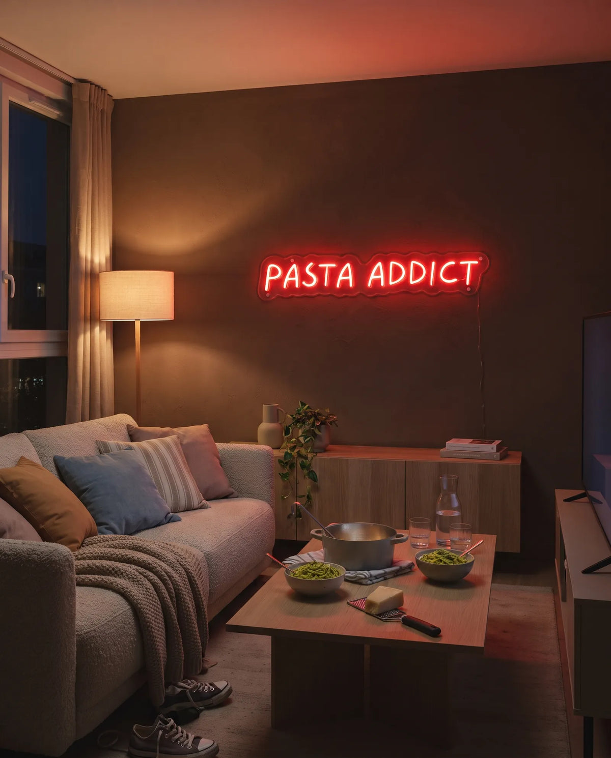 Néon LED pasta dans un salon le soir, casserole et bols de pâtes au pesto sur la table basse, canapé
