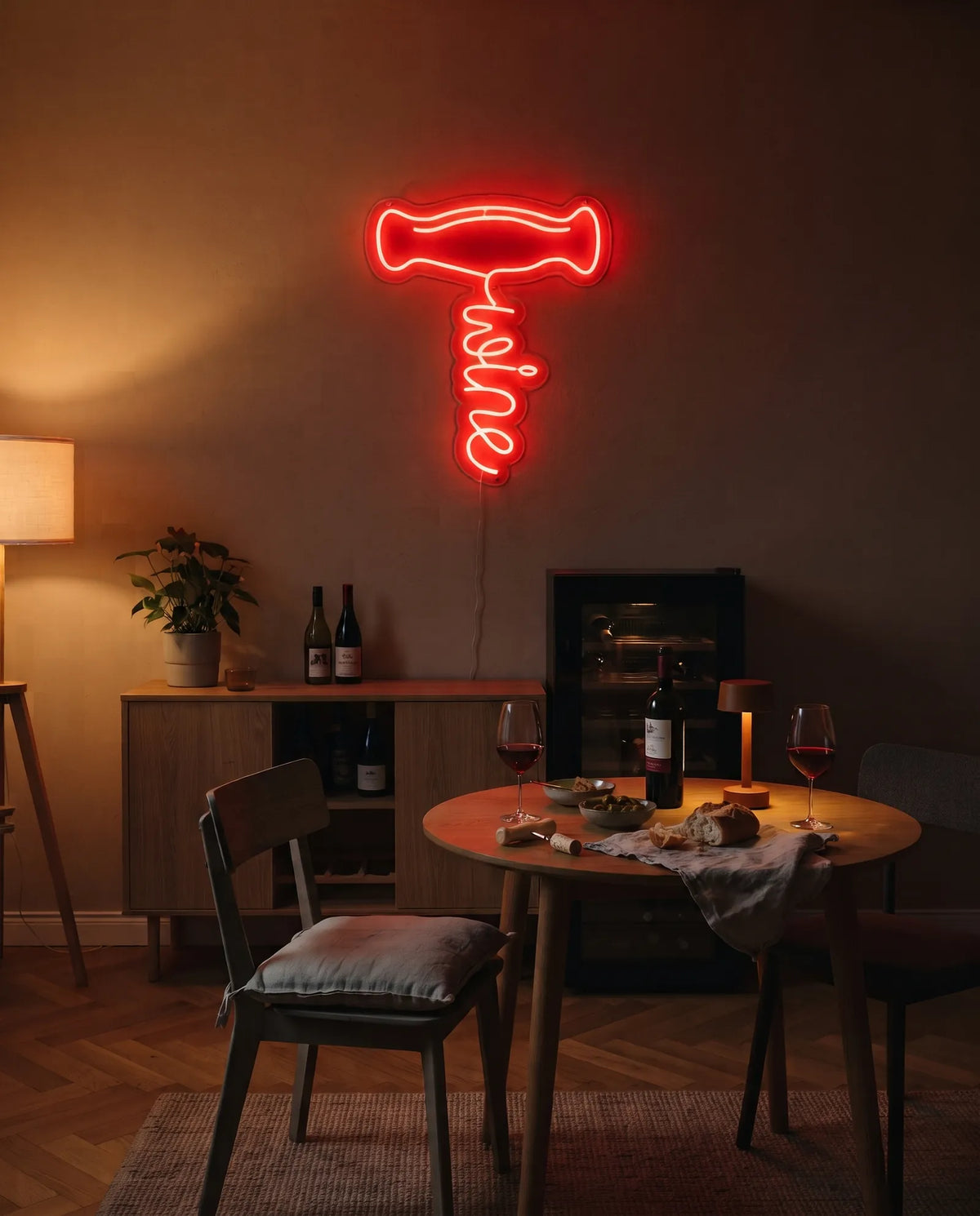 Néon tire-bouchon rouge allumé au-dessus d’un buffet et d’une cave à vin, table ronde avec pain, olives et deux verres